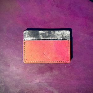 May include: A bi-color leather card holder. The top section is black, and the bottom section is a gradient of orange and red. The edges are stitched with a contrasting thread. A small logo is embossed on the lower right corner.