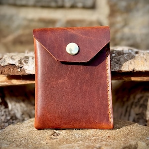 May include: A brown leather wallet with a flap closure and a silver button. The wallet is rectangular and appears to be handmade, with visible stitching along the edges. The leather has a textured appearance.