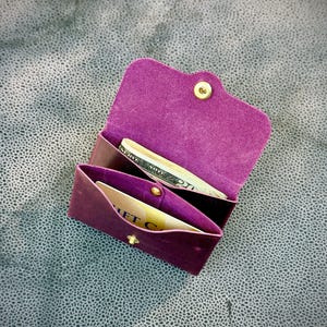 May include: A small, open, purple leather wallet with a gold snap closure. The wallet contains a gift card and folded bills. The interior is a lighter shade of purple. The wallet is on a textured gray surface.