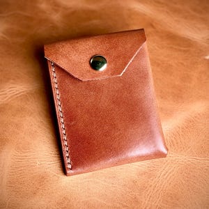 May include: A tan leather card holder featuring a triangular flap closure secured with a single brass-colored metal snap button. The side edge is finished with visible, thick, light-colored stitching along the vertical seam.