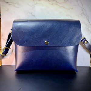 Può includere: Una borsa a tracolla in pelle blu navy con chiusura a patta e bottone color oro. La borsa ha una lunga tracolla regolabile con finiture color oro. La borsa è rettangolare e sembra fatta a mano.