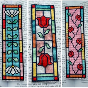 May include: Three rectangular cross-stitch bookmarks featuring stained glass window designs. The first shows a white flower on a blue background, the second features two red tulips on a pink and white grid, and the third displays red roses on a pink background.