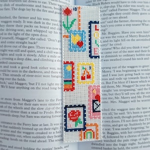 以下が含まれることがあります： チューリップ、さくらんぼ、虹など、さまざまなデザインが刺繍された正方形が特徴の手作りのブックマーク。 ブックマークは白い生地でできており、開いた本の上に置かれています。
