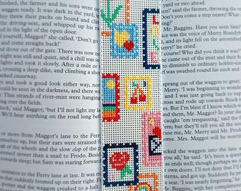 May include: Embroidered bookmark with colourful squares depicting a tulip, cherries, and a rainbow. The bookmark is on white fabric and is placed on an open book. The bookmark is approximately 18 cm long.
