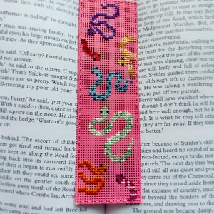 May include: A pink cross-stitch bookmark with embroidered snakes in various colours. The bookmark is rectangular and has a grid-like texture, with snakes in purple, yellow, blue, green, orange, and pink.