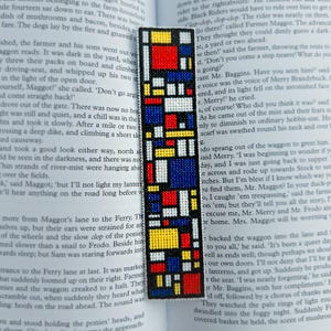 Peut inclure: Un signet fait à la main avec un motif géométrique coloré. Le signet présente un motif de carrés et de rectangles rouges, jaunes, bleus et blancs, ressemblant à une composition d'art moderne. Il est placé sur une page de livre.