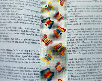 May include: A hand-stitched bookmark with a repeating pattern of embroidered butterflies. The butterflies are in shades of red, yellow, blue, and green, set against a white background with a grid pattern. The bookmark is rectangular and has a green border.