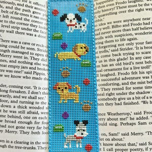 May include: A blue bookmark with a cross-stitch design featuring multiple dogs in various poses, along with colorful paw prints and shapes. The bookmark is rectangular and has a stitched border.