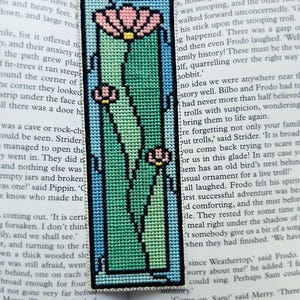 May include: Embroidered bookmark with a floral design. The bookmark features a rectangular shape with a light blue border, enclosing a green and pink floral pattern. The bookmark is made of fabric and is placed on top of a book.