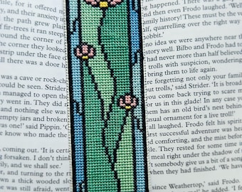 May include: Embroidered bookmark with a floral design. The bookmark features a rectangular shape with a light blue border, enclosing a green and pink floral pattern. The bookmark is made of fabric and is placed on top of a book.