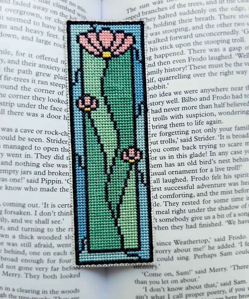 May include: Embroidered bookmark with a floral design. The bookmark features a rectangular shape with a light blue border, enclosing a green and pink floral pattern. The bookmark is made of fabric and is placed on top of a book.