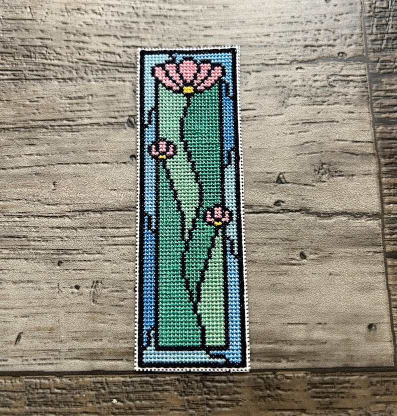 May include: A handmade bookmark with a cross-stitch floral design. The rectangular bookmark has a white border. The design features pink flowers with yellow centres, green leaves, and a blue background.