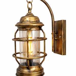 Könnte beinhalten: Eine Wandleuchte im Laternenstil mit einer antiken Messingoberfläche in Used-Optik. Das Design umfasst einen zylindrischen Schirm aus klarem Glas, der von einem Metallkäfig umschlossen und an einem geschwungenen Arm an einer rechteckigen Wandplatte befestigt ist.