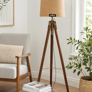 May include: A floor lamp with a brown wooden tripod base and brass hardware, featuring a cylindrical tan fabric lampshade with a black Philips brand label attached to the front. A thin black power cord hangs down from the center of the lamp.