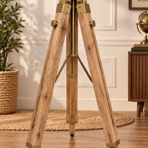 May include: A floor-standing tripod stand constructed from light brown wood with visible grain patterns. The tripod features metallic brass-toned hardware, including adjustable knobs, corner brackets, and a central mounting head at the top.