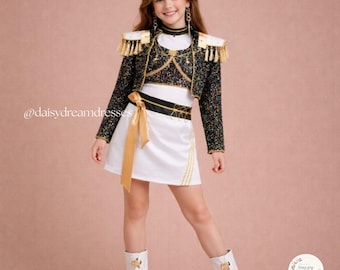 May include: A black sequinned jacket with gold fringe epaulettes worn over a white top and a white skirt with a gold sash. The outfit features gold trim and decorative chains. The text @daisydreamdresses and Daisy Dream Dresses logo are visible.