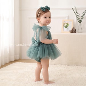 May include: A sage green tulle romper featuring sheer ruffled shoulder straps, a large bow detail on the back, and a matching headband with a bow. The garment is layered with soft tulle fabric and includes the text @dreamdaisydresses.