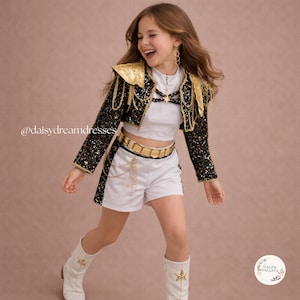 Könnte beinhalten: Ein Set aus weißem bauchfreiem Oberteil und weißer Shorts, kombiniert mit einer schwarzen Pailletten-Bolerojacke mit goldenen Schulterpolstern, Goldketten und einem Goldgürtel. Das Outfit umfasst weiße Stiefel mit goldenen Sternen und den Text @daisydreamdresses.