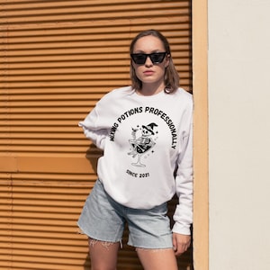 May include: White sweatshirt with a black graphic of a skeleton in a martini glass, wearing a witch hat. The text reads "Mixing Potions Professionally Since 2021". The person is wearing denim shorts and sunglasses.