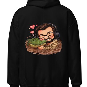 Pode incluir: Moletom preto com uma ilustração de desenho animado de uma pessoa usando óculos e fones de ouvido, abraçando um jacaré verde em uma poça marrom. Corações rosa flutuam acima. O moletom é feito de um material macio.