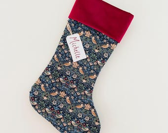 May include: A Christmas stocking with a dark blue floral pattern featuring birds and foliage. The top of the stocking is a deep red velvet. A tag with the name "Michelle" is attached.