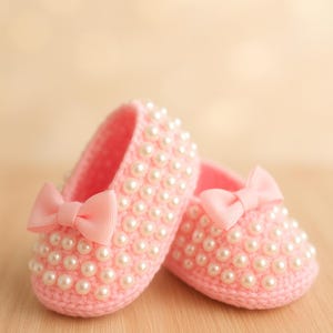 May include: A pair of pink baby shoes, decorated with faux pearls and a bow. The shoes are made of a soft, knitted material. The background is a light, blurred colour.