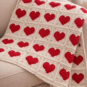 Può includere: Una coperta all'uncinetto color crema con un motivo ripetuto di cuori rossi. La coperta è drappeggiata su un divano beige. Le parole "Blanket Crochet Pattern" sono stampate in alto e in basso nell'immagine.