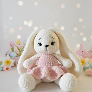 May include: A handmade white crochet bunny wearing a pink dress. The bunny has long, floppy ears and black eyes. The background includes Easter decorations like eggs and flowers, suggesting a spring theme.