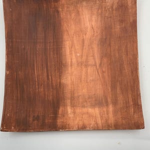 Op de afbeelding: Een vierkante, handgemaakte, bruine keramische plaat met een licht gebogen rand. De plaat heeft een getextureerd oppervlak met zichtbare penseelstreken, waardoor het een rustieke uitstraling heeft. De kleur is een warm, aards bruin.