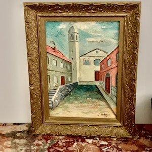 Op de afbeelding: Olieverfschilderij van een Europees stadsplein met een kerk en een klokkentoren. De gebouwen zijn geschilderd in tinten rood, grijs en wit, met een blauwe lucht. Het schilderij zit in een gouden, sierlijke lijst.