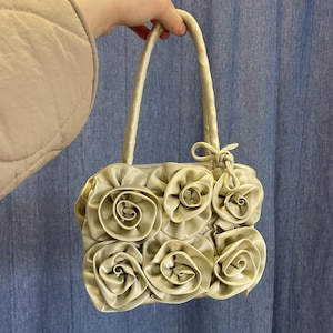 Op de afbeelding: Een lichtgroene handtas met een bloemenontwerp. De tas heeft meerdere stoffen rozen aan de voorkant en een paar handvatten. De tas wordt door een persoon voor een blauwe achtergrond vastgehouden.