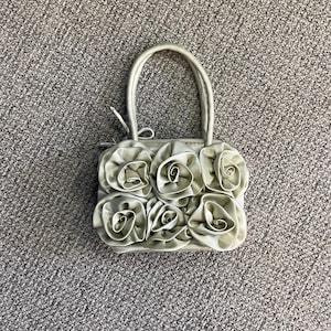 Op de afbeelding: Zilverkleurige handtas met een handvat, versierd met grote stoffen rozen. De tas heeft een rechthoekige vorm en een satijnachtige textuur, ideaal voor speciale gelegenheden.