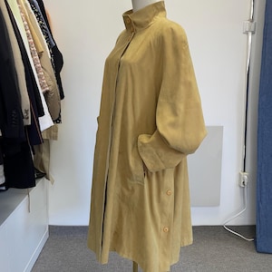 May include: A mustard yellow coat with a high collar, long sleeves, and a button-front closure. The coat features a smooth, matte texture and side pockets, displayed on a mannequin with a wire mesh head form in a side profile view.