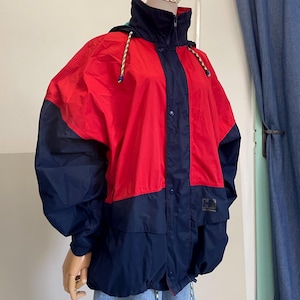 May include: A Helly Hansen windbreaker jacket featuring a color-blocked design in red and navy blue. The jacket includes a high collar, a front zipper closure with a snap-button placket, and a hood with adjustable drawstrings. The brand logo is visible.