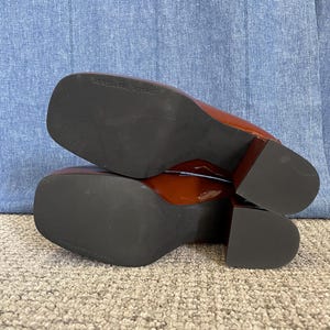 May include: Pair of glossy, reddish-brown heeled shoes with square toes. The soles are black, and the heels are a block style. The shoes are resting on a light-colored carpet.