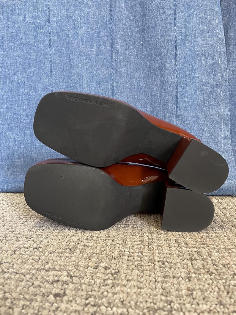 May include: Pair of glossy, reddish-brown heeled shoes with square toes. The soles are black, and the heels are a block style. The shoes are resting on a light-colored carpet.