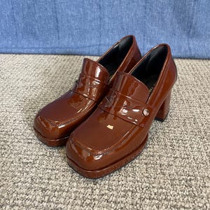 May include: A pair of brown patent leather heeled loafers. The shoes have a square toe, a strap across the top with a silver-colored button, and a chunky heel. The shoes are on a gray carpet.