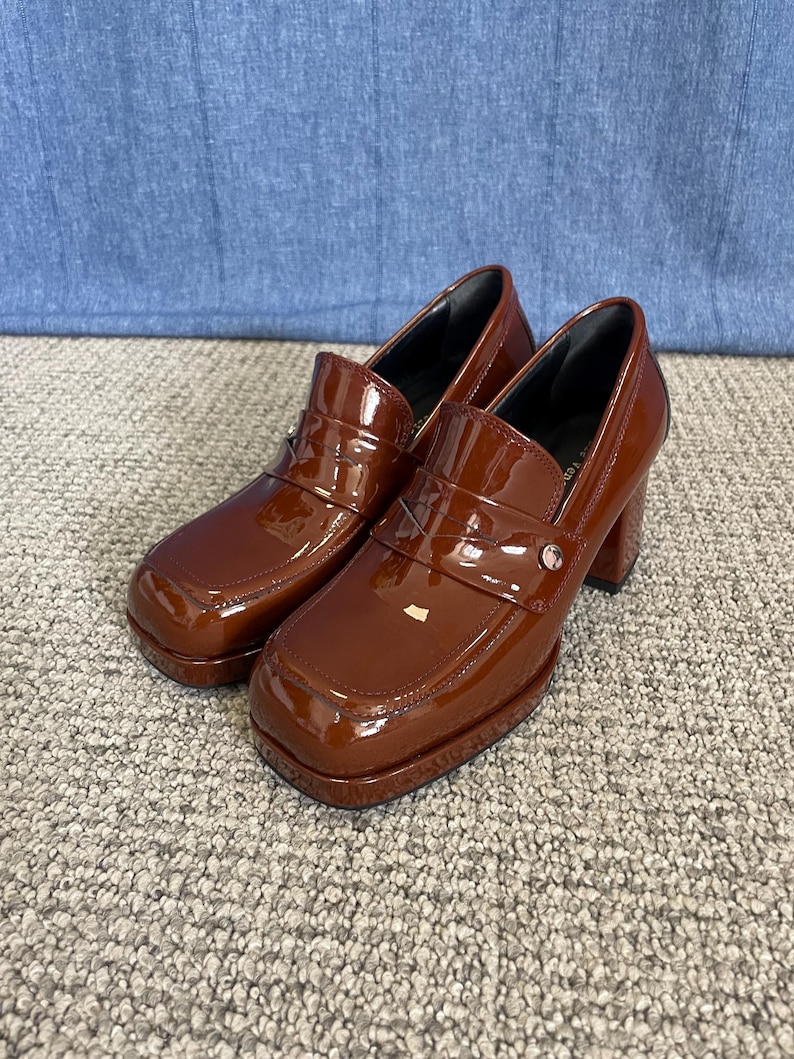 May include: A pair of brown patent leather heeled loafers. The shoes have a square toe, a strap across the top with a silver-colored button, and a chunky heel. The shoes are on a gray carpet.