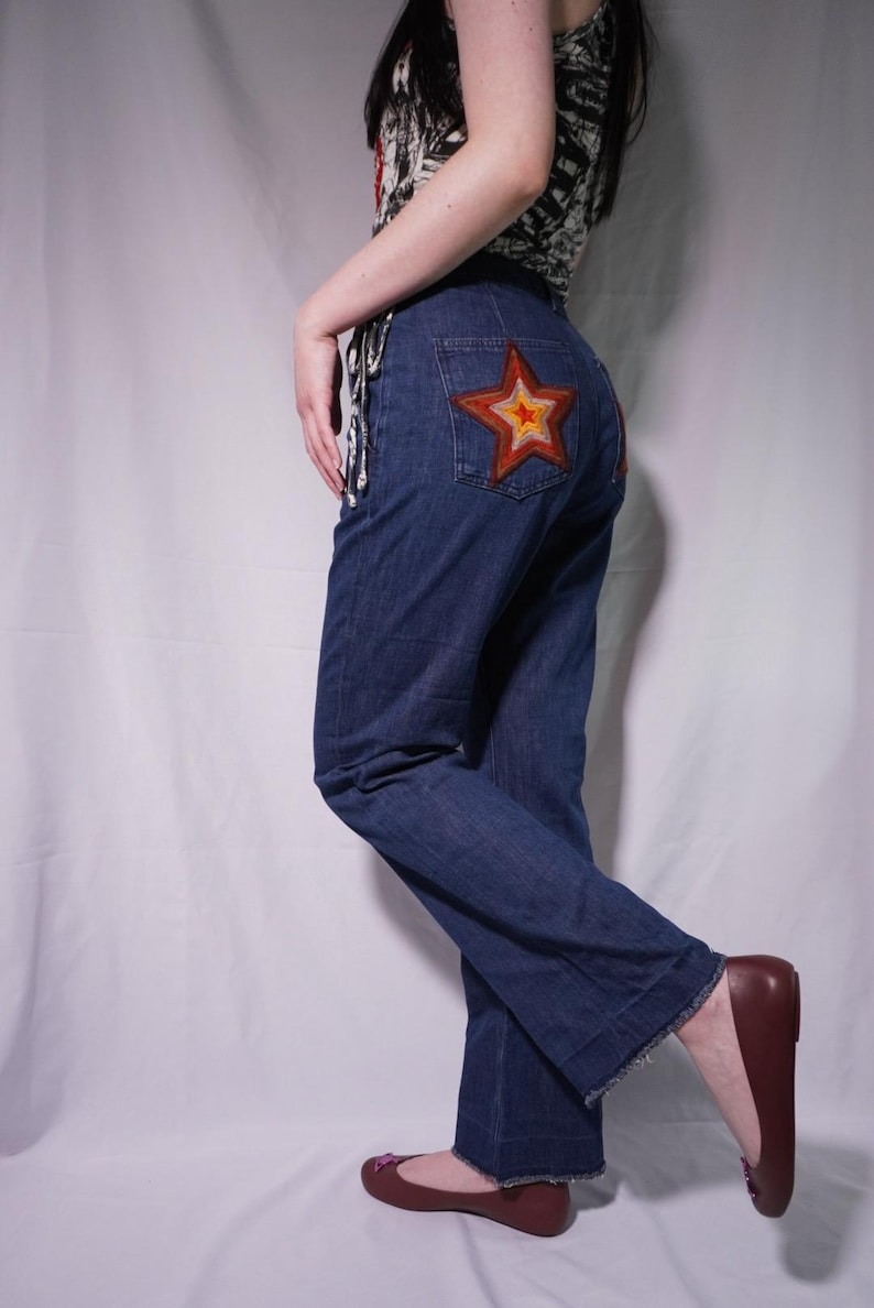 May include: Dark blue denim jeans featuring a prominent embroidered star on the back pocket. The star design consists of concentric layers in shades of brown, orange, and yellow. The jeans have a straight-leg cut with a raw, frayed hem.