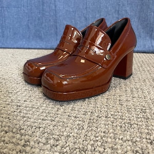 May include: A pair of brown platform heeled loafers with a glossy finish. The shoes have a square toe, a strap across the top, and a chunky heel. The shoes are displayed on a neutral carpet.