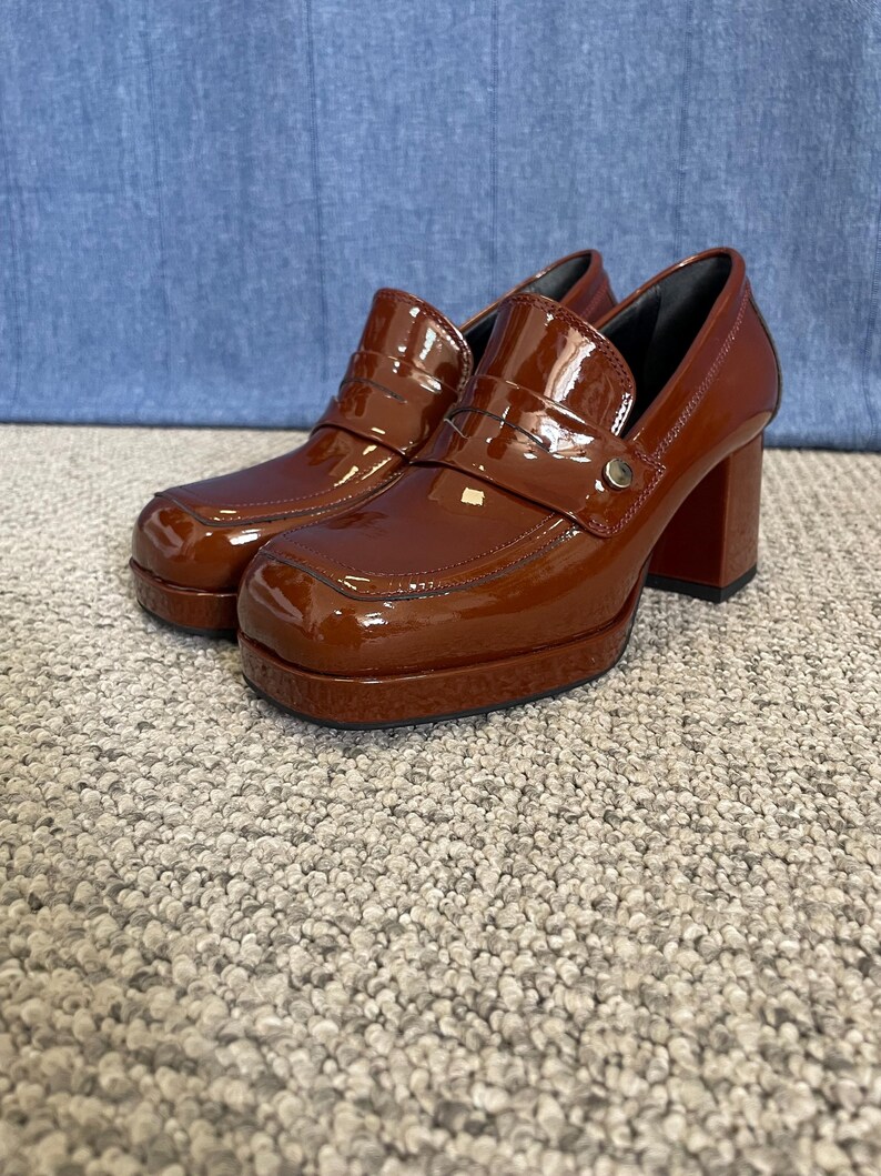 May include: A pair of brown platform heeled loafers with a glossy finish. The shoes have a square toe, a strap across the top, and a chunky heel. The shoes are displayed on a neutral carpet.