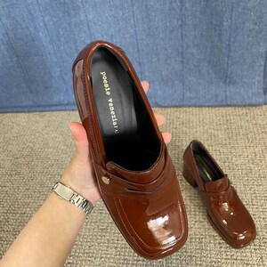 May include: A pair of glossy, brown leather loafers with a square toe and small heel. The shoes have a decorative strap and a gold-colored button. The brand name "poesie veneziane" is printed on the insole.