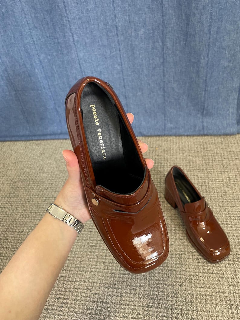 May include: A pair of glossy, brown leather loafers with a square toe and small heel. The shoes have a decorative strap and a gold-colored button. The brand name "poesie veneziane" is printed on the insole.