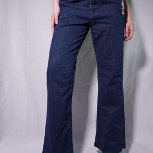 May include: Dark blue denim jeans with a slight flare at the hem. The jeans have a classic five-pocket design and a button and zipper closure. The model is wearing dark red ballet flats.