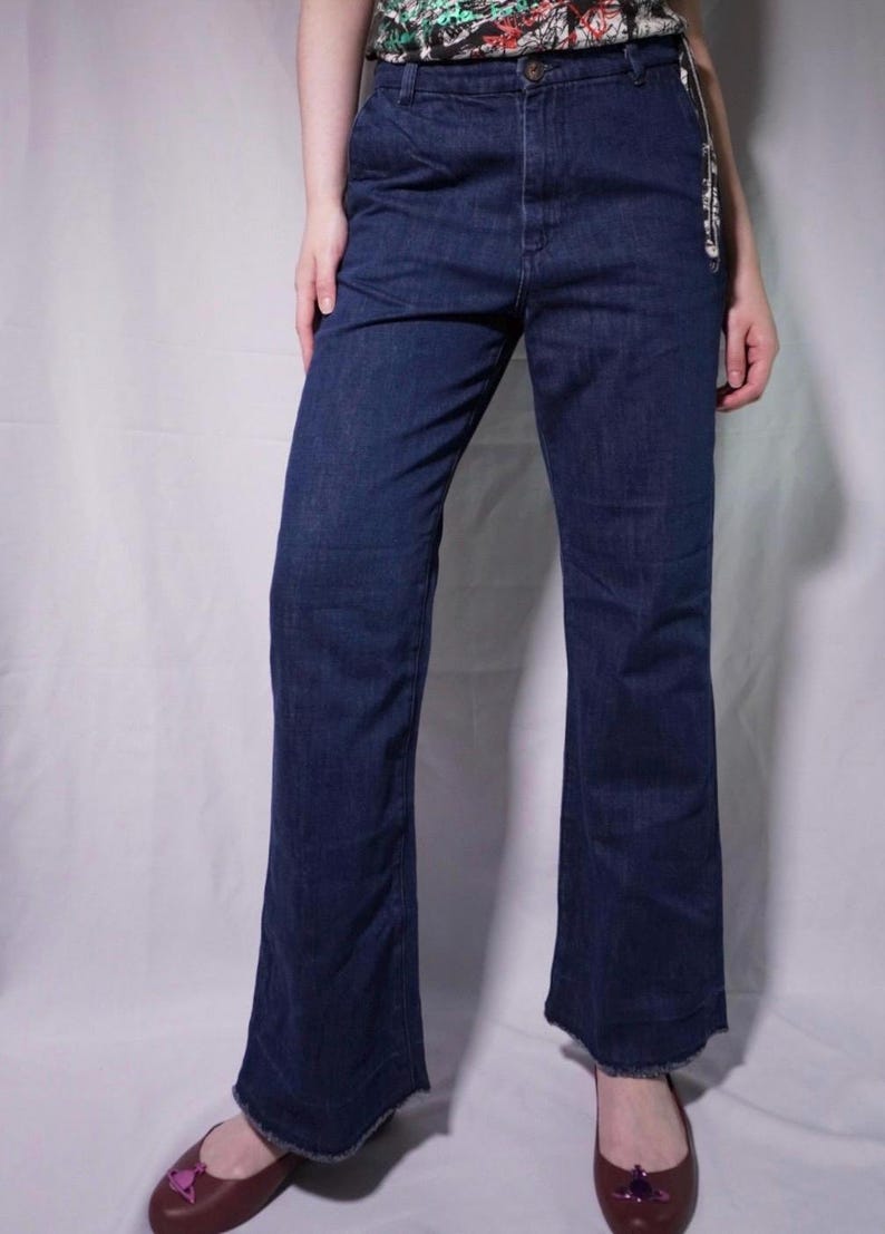 May include: Dark blue denim jeans with a slight flare at the hem. The jeans have a classic five-pocket design and a button and zipper closure. The model is wearing dark red ballet flats.