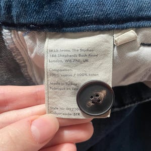 May include: A dark blue denim garment featuring an interior fabric label with the text M.i.h Jeans, The Studios, 186 Shepherds Bush Road, London, W6 7NL, UK, Composition 100% cotton, Made in Turkey, Style Nr D2210, and a dark brown button.