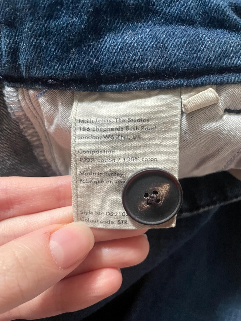 May include: A dark blue denim garment featuring an interior fabric label with the text M.i.h Jeans, The Studios, 186 Shepherds Bush Road, London, W6 7NL, UK, Composition 100% cotton, Made in Turkey, Style Nr D2210, and a dark brown button.