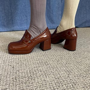 May include: Brown platform loafers with a glossy finish. The shoes have a chunky heel and a classic penny loafer design. The image shows the shoes paired with socks, one pair is taupe ribbed and the other is cream cable knit.