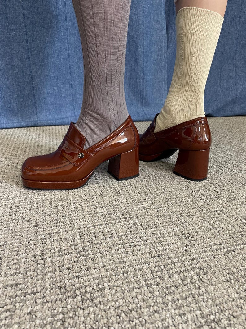 May include: Brown platform loafers with a glossy finish. The shoes have a chunky heel and a classic penny loafer design. The image shows the shoes paired with socks, one pair is taupe ribbed and the other is cream cable knit.