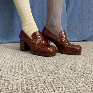 May include: Brown patent leather platform loafers with a chunky heel. The shoes have a classic penny loafer design with a rounded toe and a small metal detail. The image shows the shoes from the ankles down, with beige and brown socks.